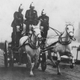 A black and white photo of firefighters rushing to an emergency on a horse-drawn fire truck.